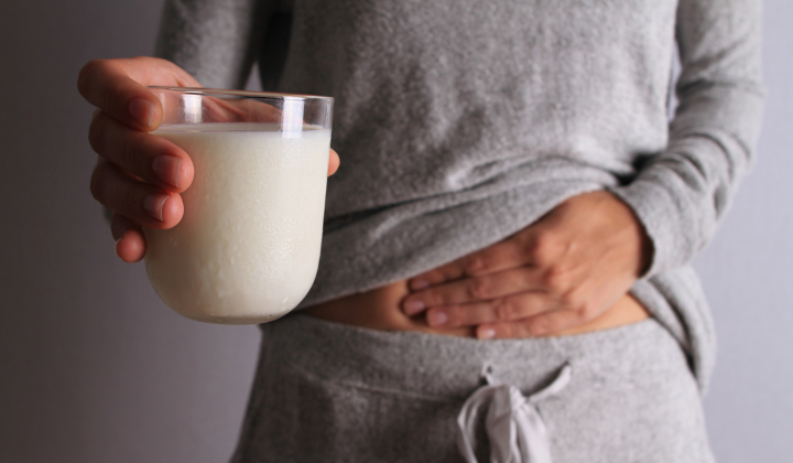 Milchunverträglichkeit Frau traegt einen bequemen, grauen Traineranzug und haelt mit ihrer rechten Hand vor Schmerzen ihren Bauch mit der linken Hand haelt sie ein Glas Milch.